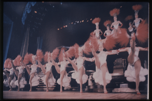 Color view of show girls in white costumes and red feather headdresses.