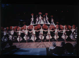 Color view of show girls in white costumes and red feather headdresses.