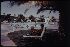 Color view of a mother and daughter lounging by a hotel pool that is surrounded by palm trees.