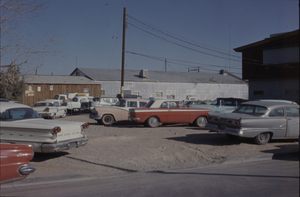 Color view of an unpaved parking lot with several buildings visible in the background.