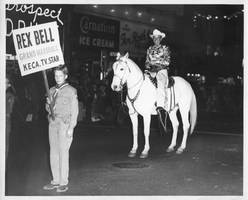 Rex Bell (George Francis Beldam) on horseback in a parade.  Boy scout in front of him is holding a sign that states: Rex Bell Grand Marshall KECA.TV.Star. Copyright notice on the back of image: Miller's Photo Service, 1644 No. Vermont Ave., Hollywood, California