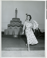 Photograph of a woman standing in front of a giant replica birthday cake outside of the Desert Inn, Las Vegas, Nevada, April 1951