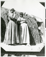 Photograph of Two Paiute women standing in front of a karnee dwelling, possibly in Pahrump Valley, Nevada, circa 1880s-1900s