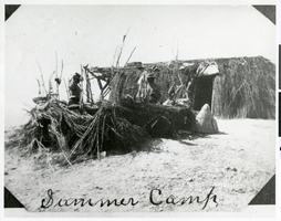 Photograph of a man and a woman standing in front of brush house, with a shade shelter on one end, possibly Ash Meadows or Pahrump, Nevada, circa 1900s-1910s