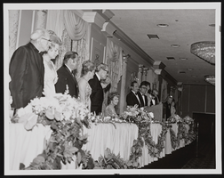Monsignor Ryan (far left), U.S. Senator Howard Cannon (third from left), U.S. Senator Alan Bible (sixth from left), Judy Bayley (seated), Gus Giuffre (standing at podium), and Governor Grant Sawyer (right of Giuffre): photographic print