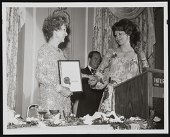 Judy Bayley accepting honroary proclamation for "Judy Bayley Day" from an unidentified woman, with Gus Giuffre standing in background: photographic print