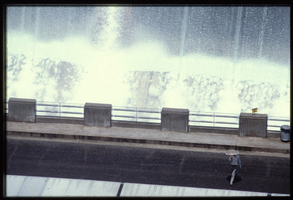 A man uses an umbrella in the water spray caused by overflow water in the Arizona spillway, looking west at Hoover Dam, Arizona: photographic slide