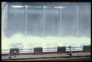 A man uses an umbrella in the water spray caused by overflow water in the Arizona spillway, looking west at Hoover Dam, Arizona: photographic slide