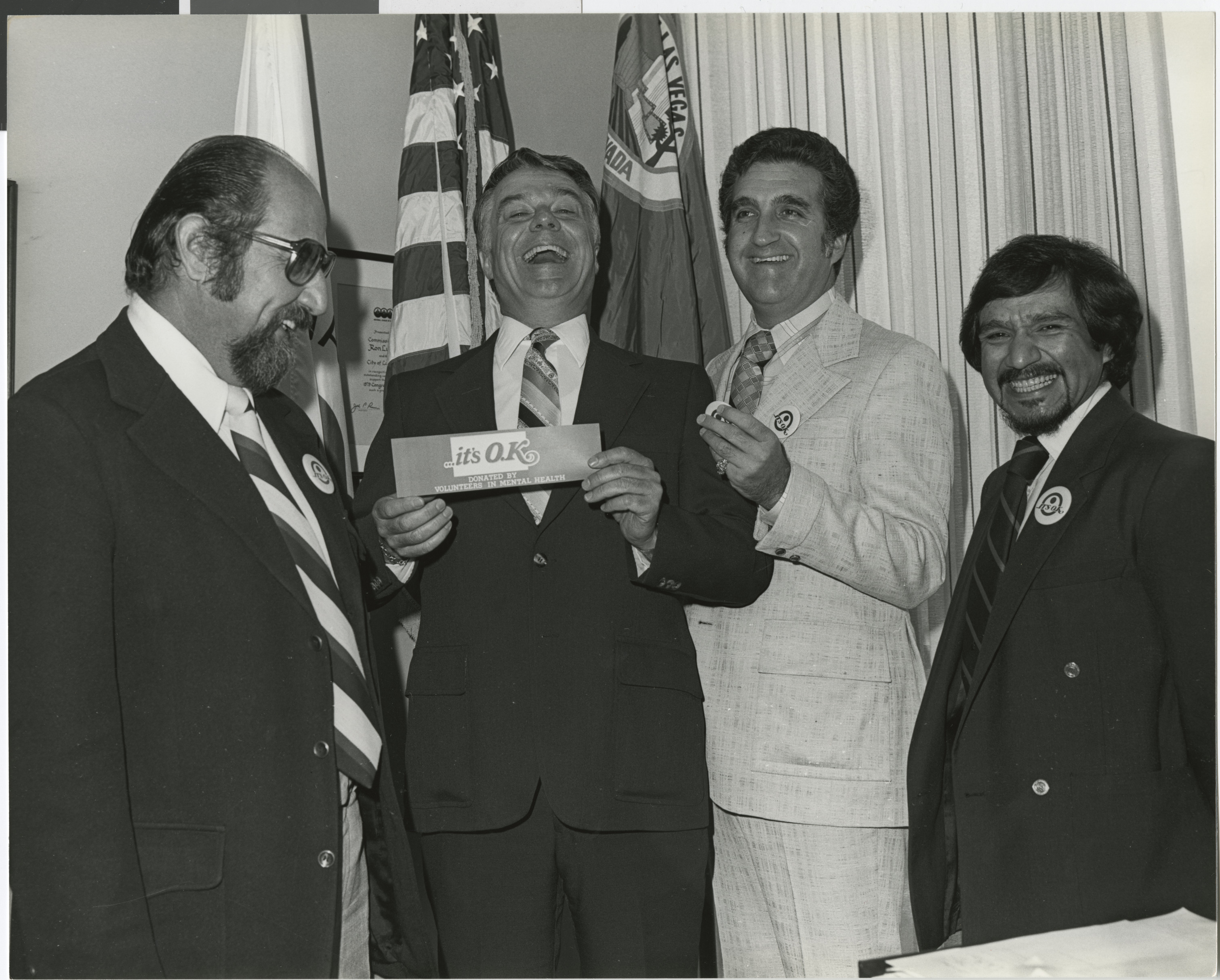 Photograph of Ron Lurie (second from right) with a group of men, including Mayor Briare holding a sticker that reads "... it's OK Donated by Voluteers in Mental Health"