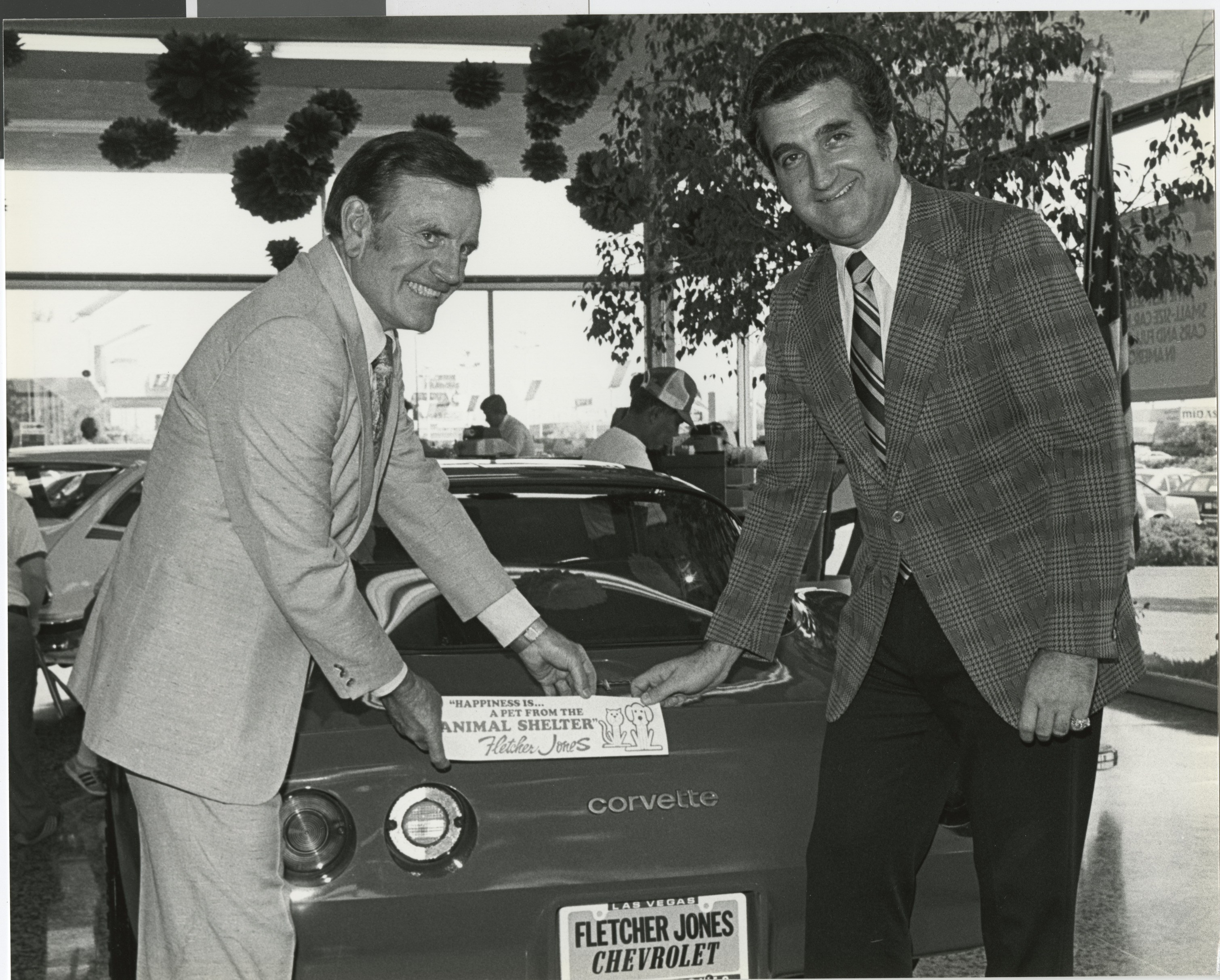 Photograph of Ron Lurie placing a bumper sticker for an animal shelter on a Corvette at Fletcher Jones car dealership